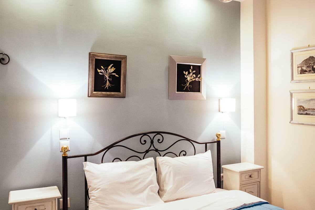 Una cama bien tendida con un colcha azul, dos mesitas de noche con lámparas, y cuadros en la pared.