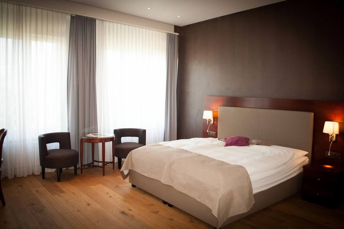 A hotel room with a large bed, two chairs, a small table, and a nightstand with a lamp.
