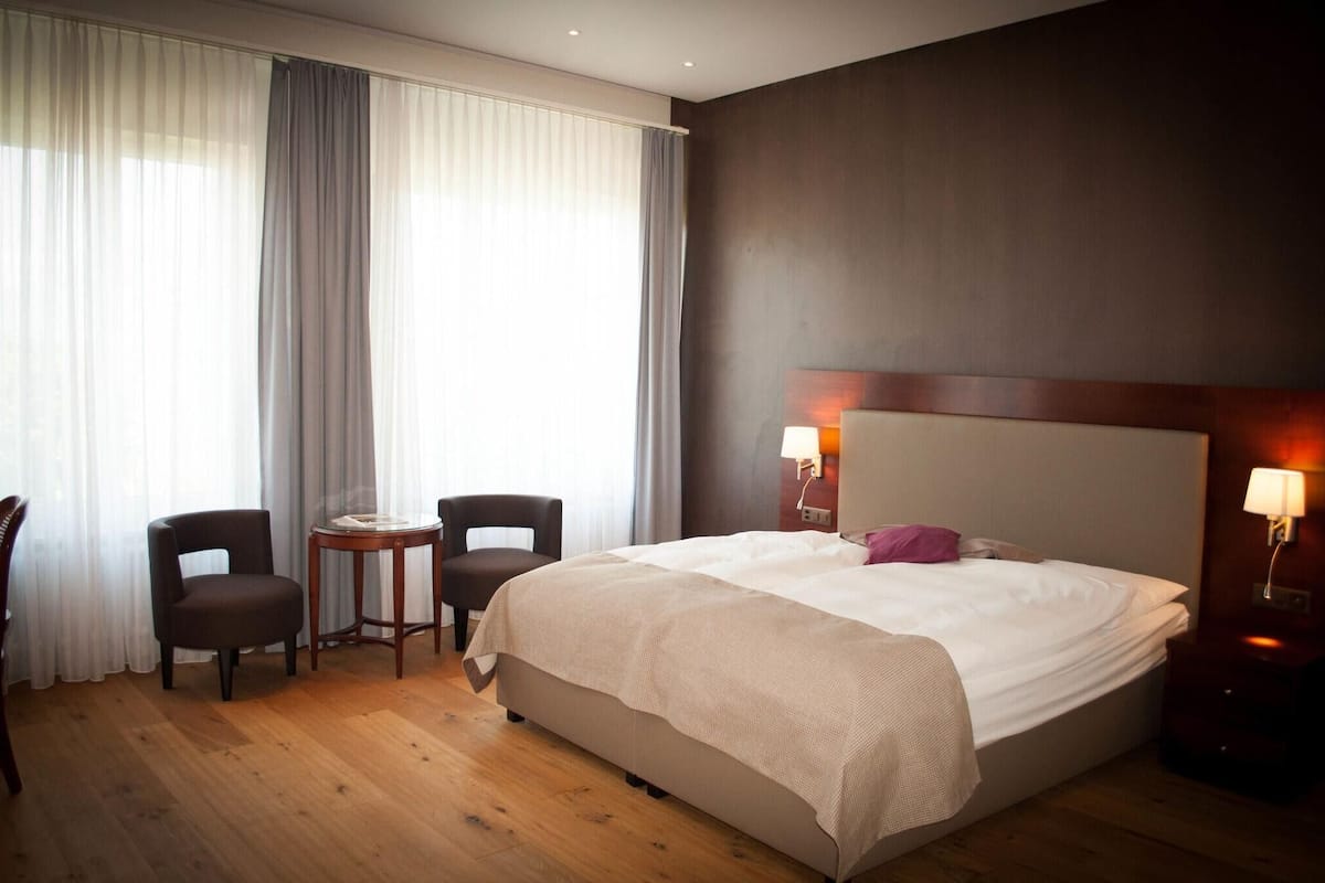 Hotellihuone, jossa on suuri sänky, kaksi tuolia, pieni pöytä ja yöpöytä, jossa on lamppu.