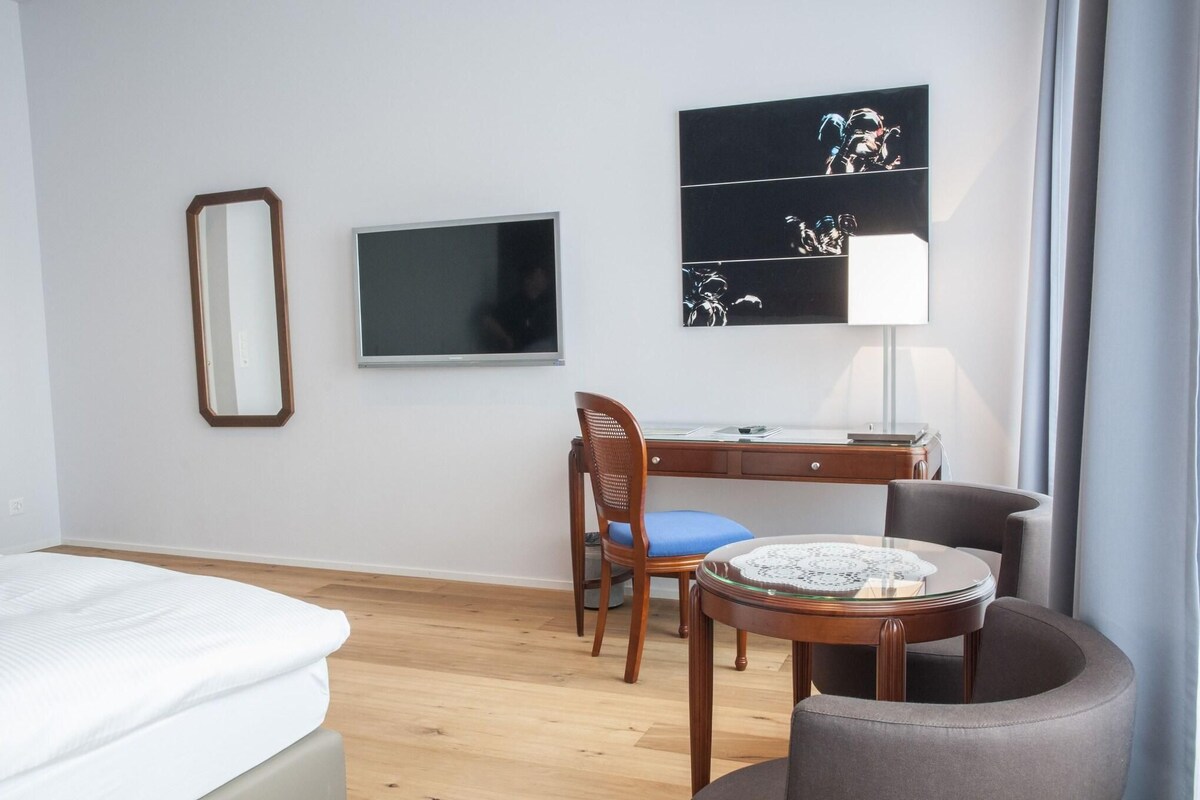 Habitación de hotel con cama, escritorio, silla, televisor, un cuadro y una lámpara.