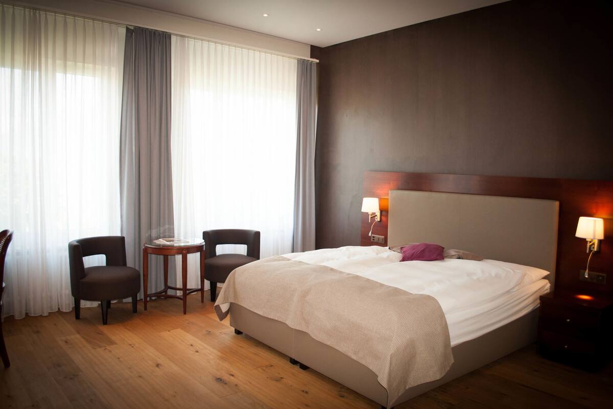 A hotel room with a large bed, two chairs, a small table, and a nightstand with a lamp.
