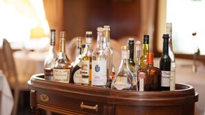 Een houten tafel met diverse alcoholische flessen, waaronder merken als Hennessy en Grappa, in een goed verlichte kamer met een onscherpe achtergrond.