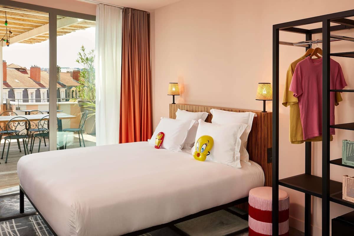 Une chambre d’hôtel comprenant un lit, une armoire, une table avec des chaises et une vue sur les bâtiments, visible à travers une porte vitrée.