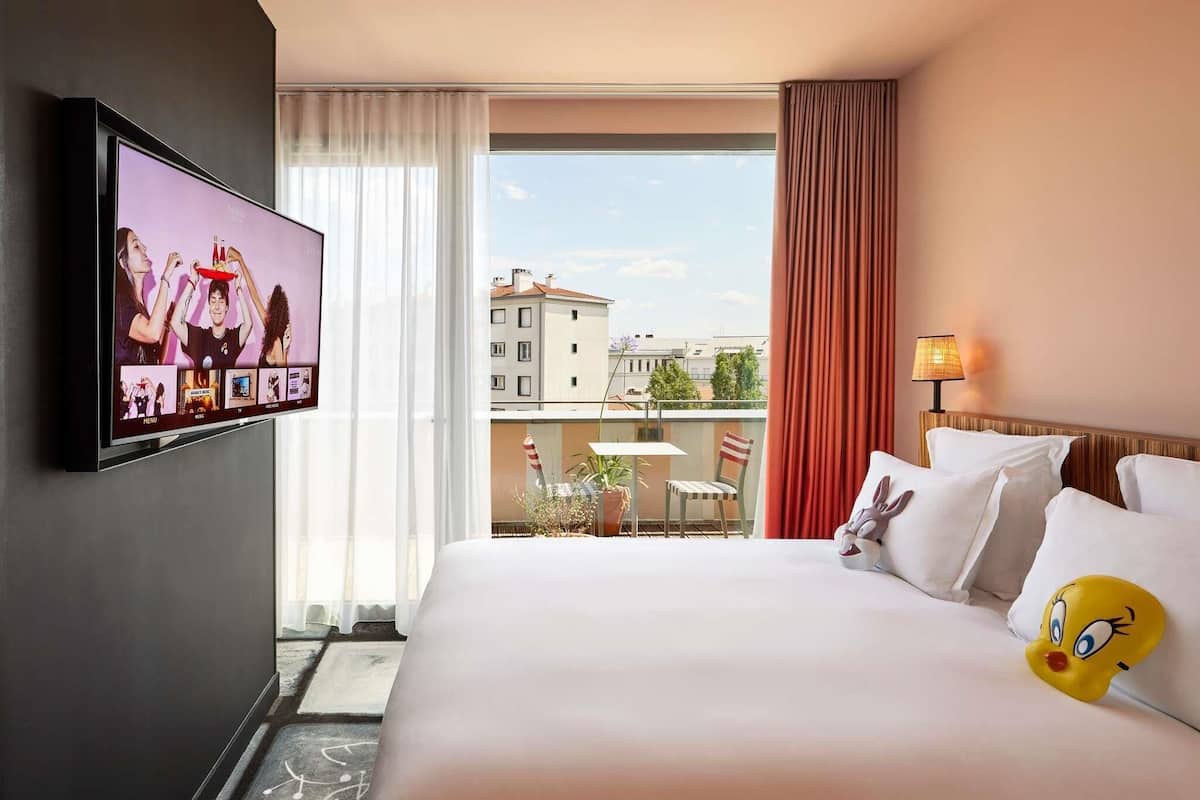 Une chambre dâhĂŽtel avec un lit, une tĂ©lĂ©vision, un balcon avec une table et des chaises, et une vue sur les bĂątiments.