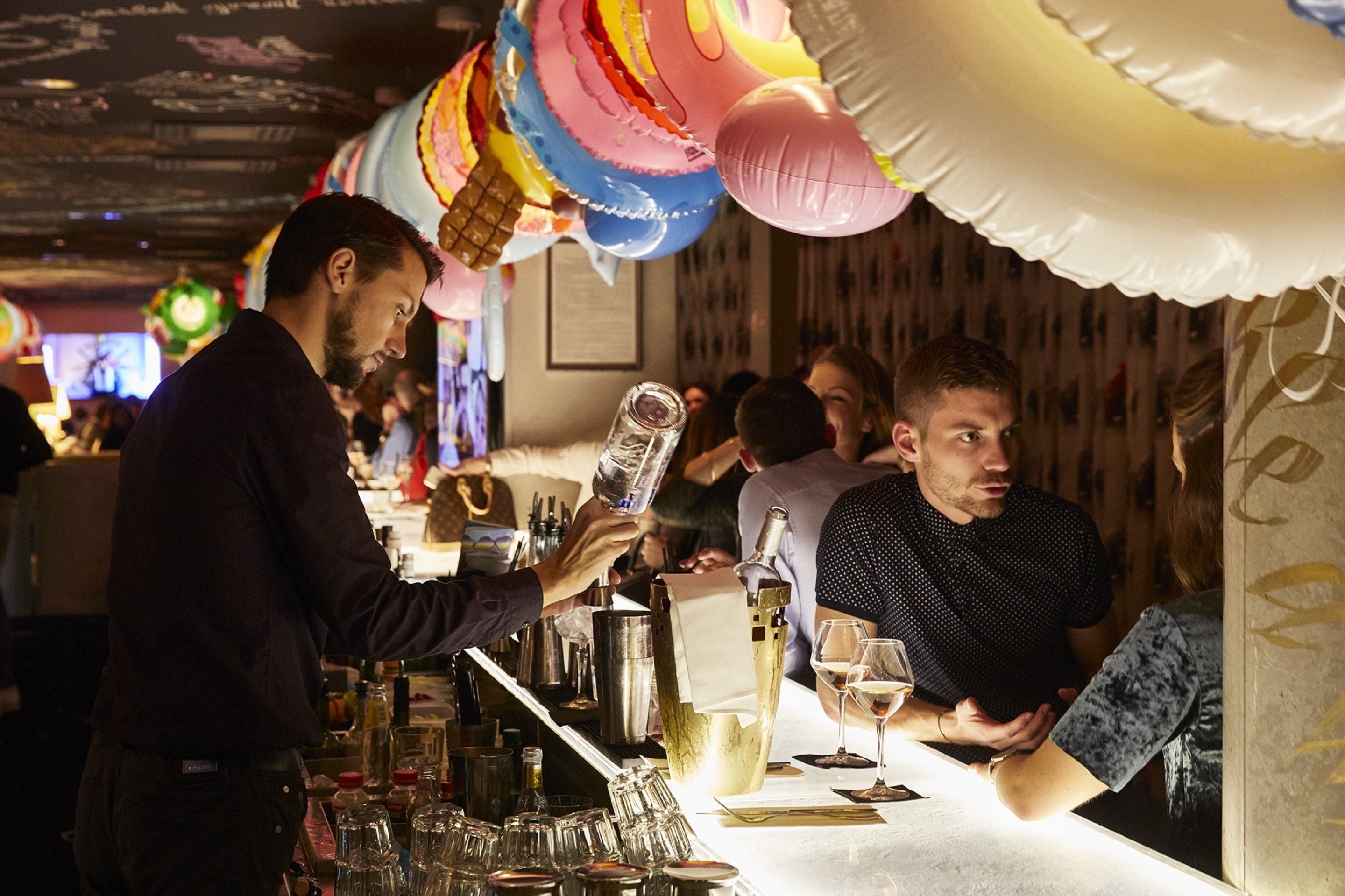 A bar area with a bartender preparing drinks, a counter with various bottles and glasses, and balloons hanging from the ceiling.