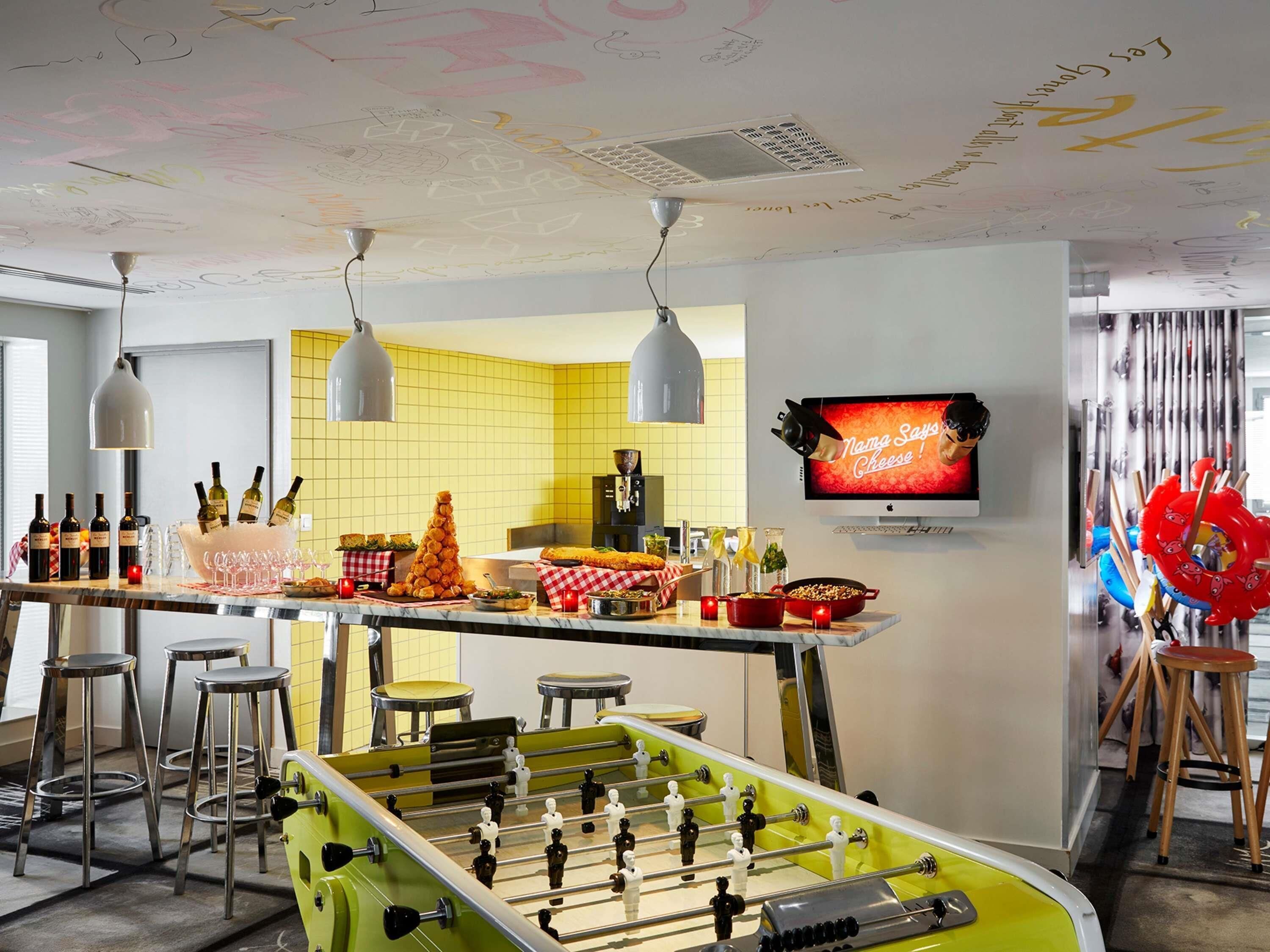 Uma área de bar com banquetas, uma mesa de futebol de mesa e uma televisão exibindo um anúncio de queijo.