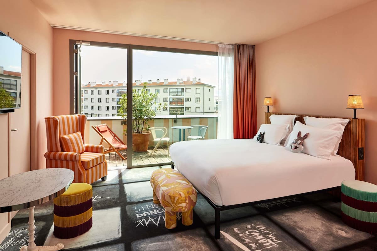 Une chambre d’hôtel avec un lit, une chaise, une petite table et un balcon donnant sur des immeubles.