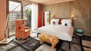 A hotel room with a large bed, a striped armchair, a small stool, a desk lamp, and a view of buildings through a window. - Mama Shelter Lyon (Lyon)