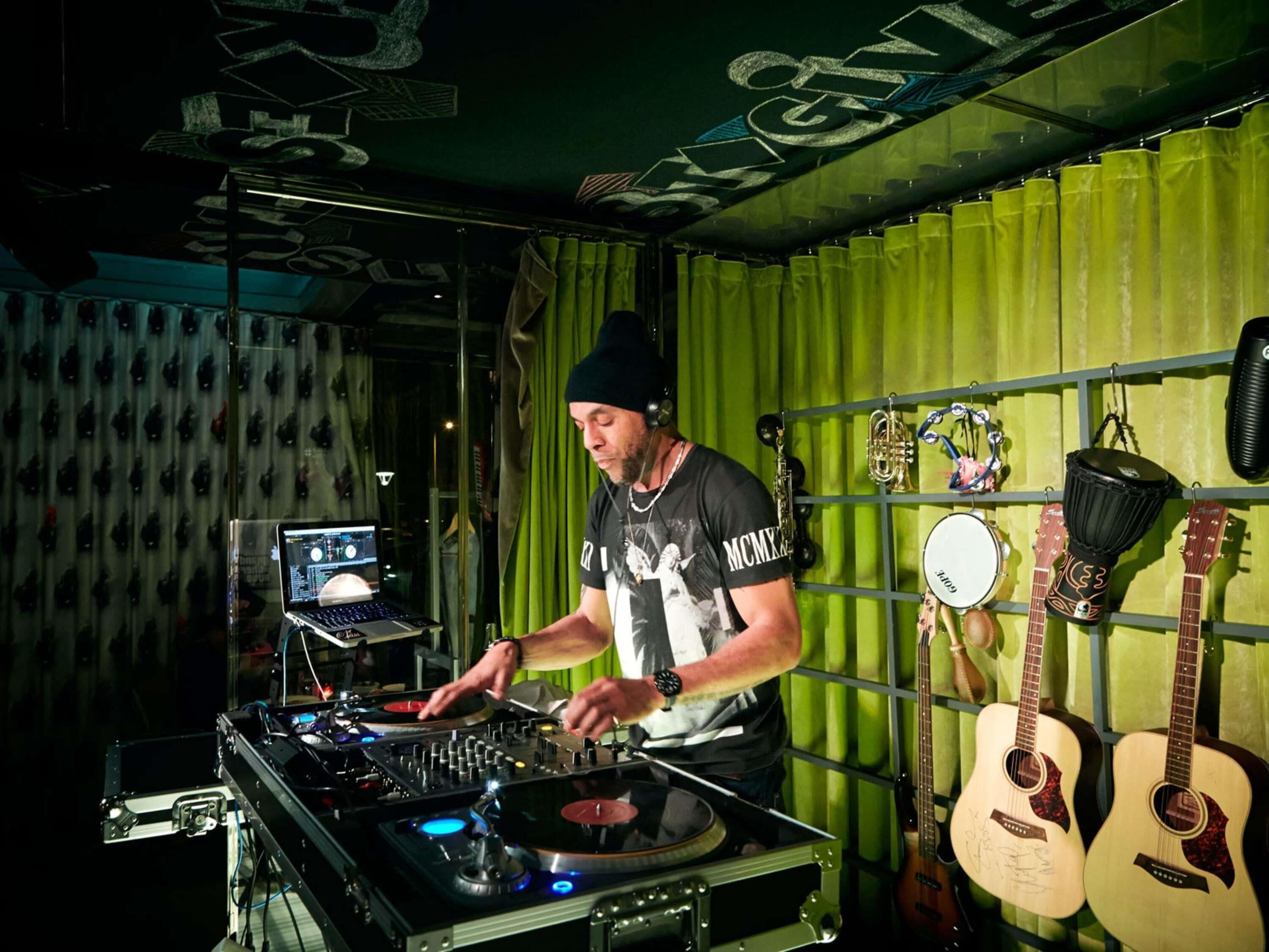 Iemand bedient DJ-apparatuur in een ruimte met een groen gordijn, een aan de muur bevestigde trommel en twee gitaren.