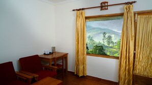 A room with a wooden floor, a wooden table, a red chair, and a view of a mountainous landscape through a window with yellow curtains. - Glenmore Resorts (Devikolam)