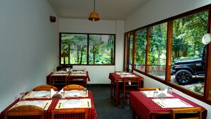 A dining area with tables set for four, large windows with a view of greenery and a vehicle outside. - Glenmore Resorts (Devikolam)