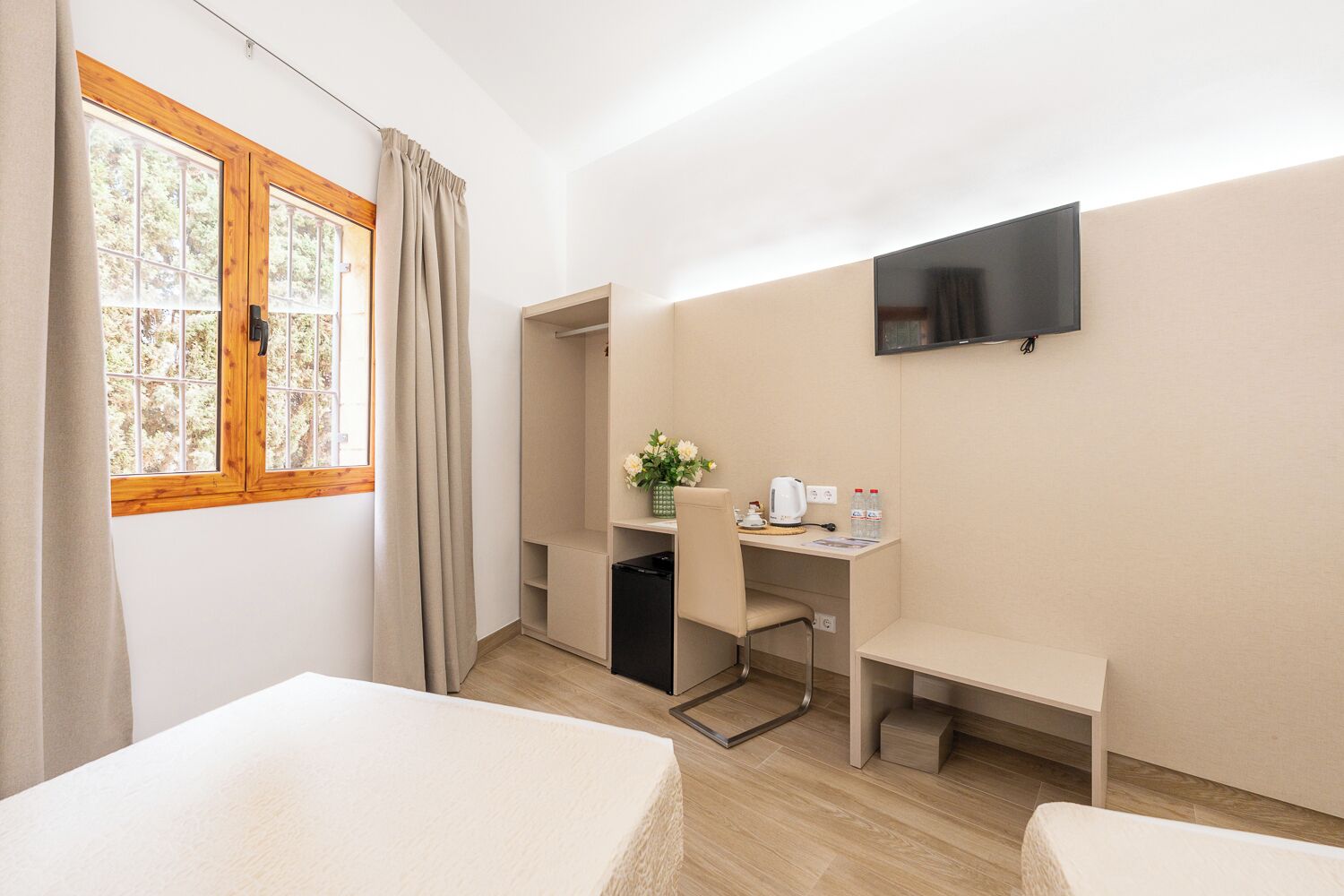 A hotel room with a bed, a desk with a chair, a TV mounted on the wall, and a window with curtains.