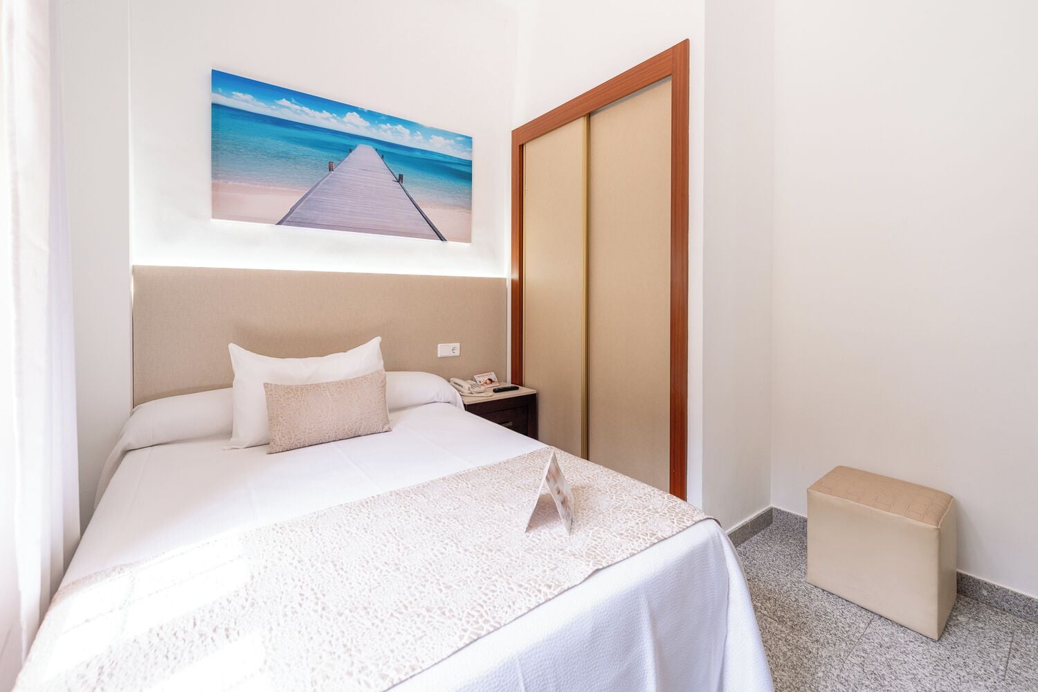 A hotel room with a bed, a nightstand, a wooden wardrobe, and a painting of a beach.
