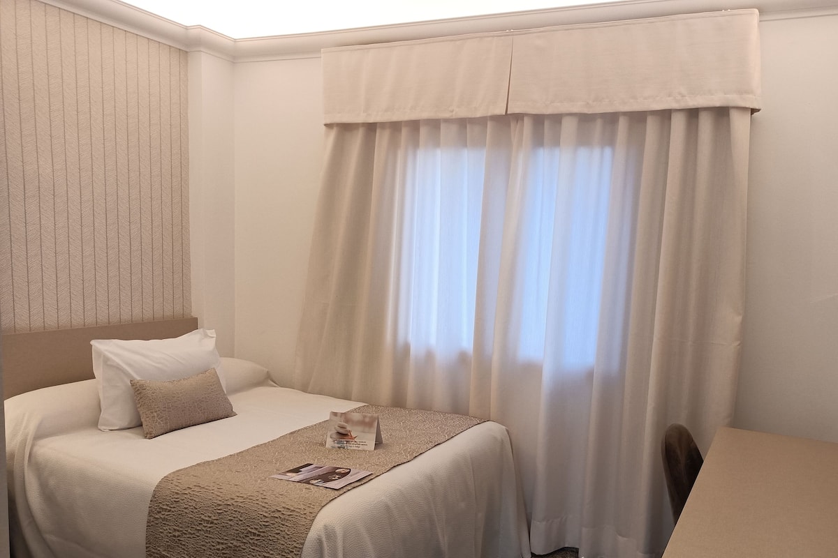 A hotel room with a bed, a flat-screen TV, and a window with sheer curtains.