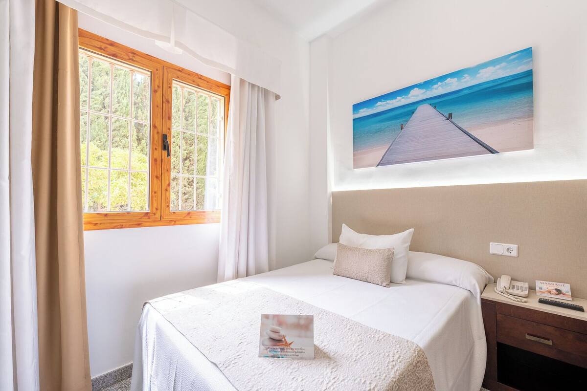 A neatly arranged bedroom with a bed, a nightstand, a painting of a beach, and a window with curtains.