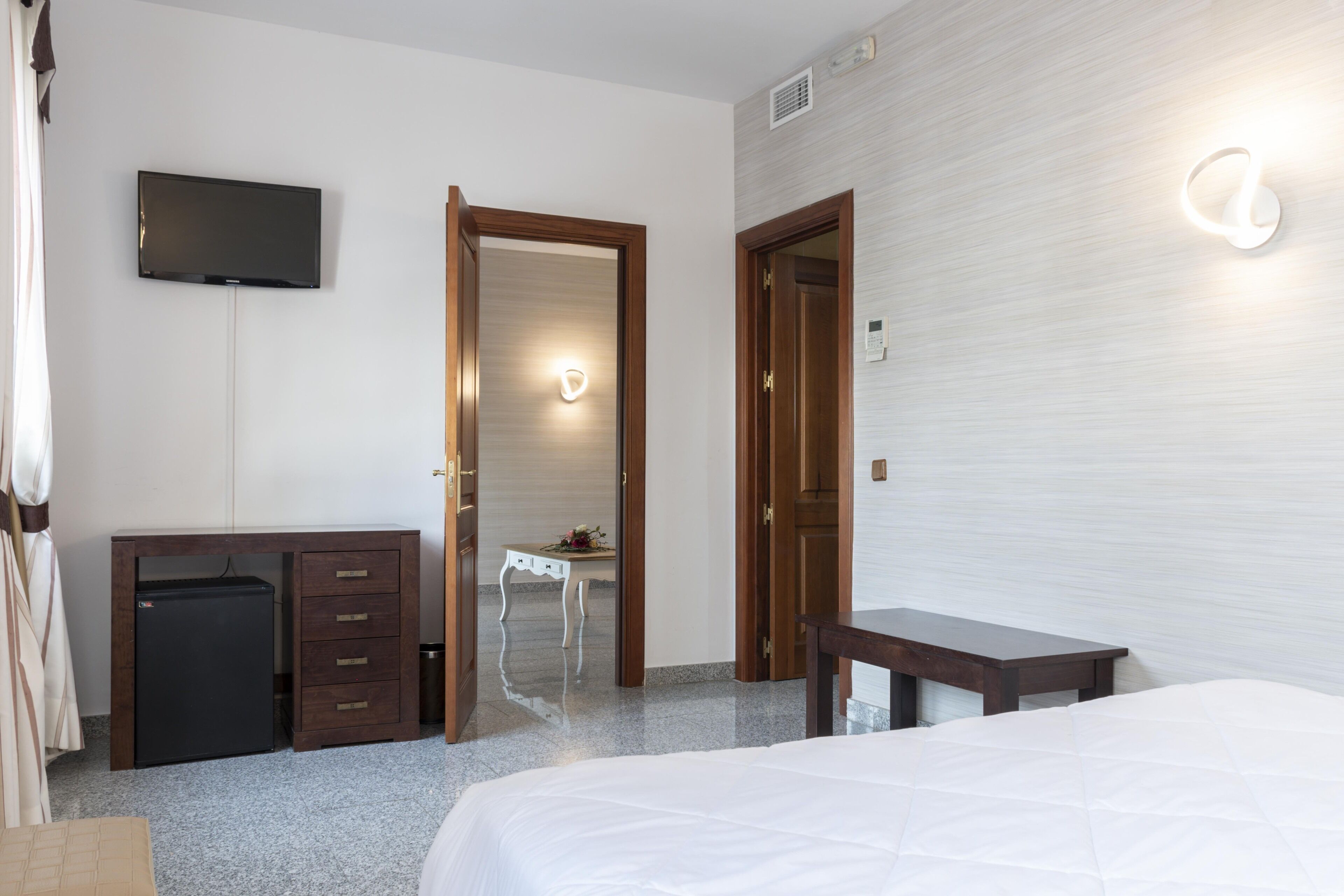 Une chambre d’hôtel équipée d’un lit, d’un bureau, d’un petit réfrigérateur, d’un téléviseur fixé au mur et d’une salle de bain visible à travers une porte ouverte.