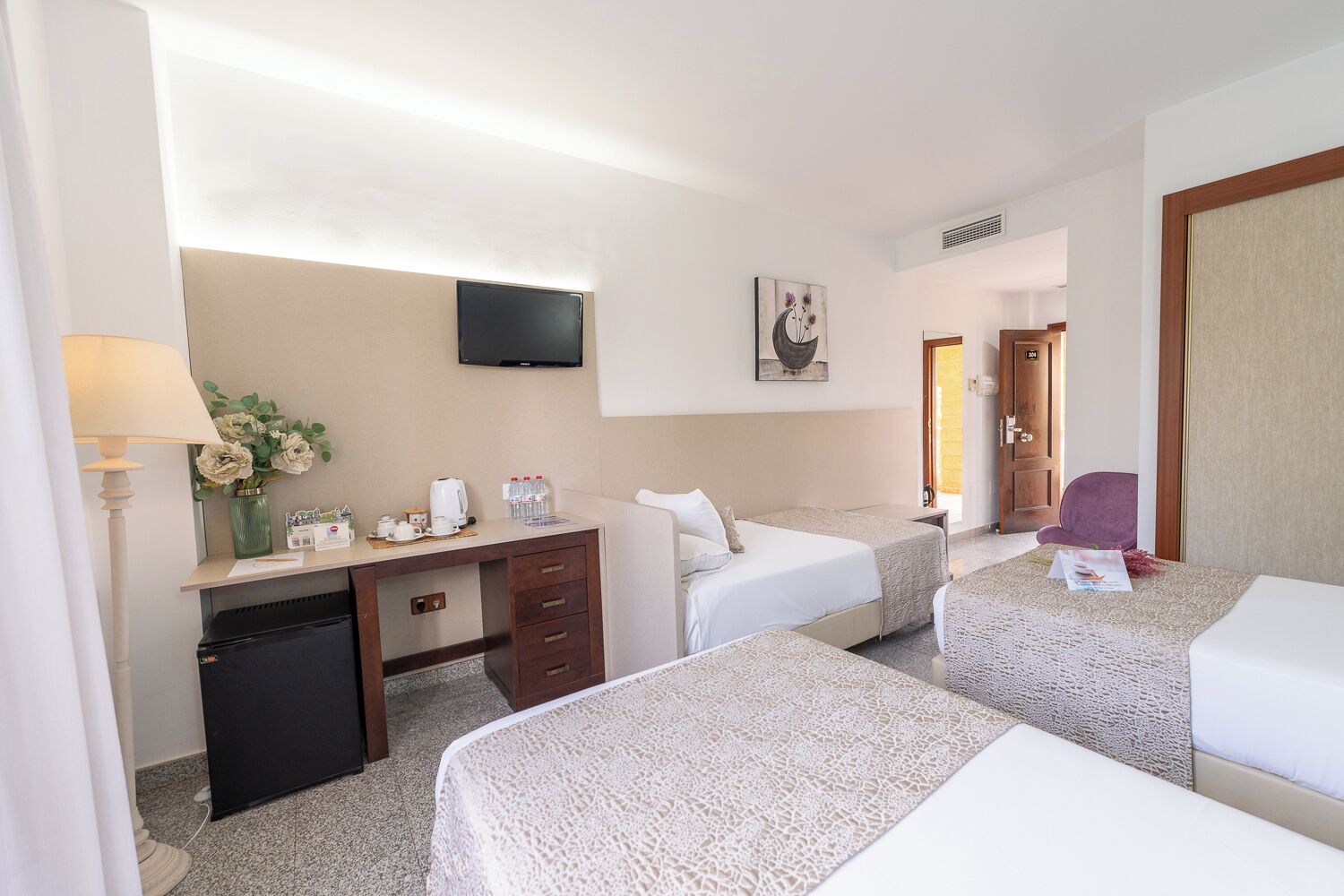 Habitación de hotel con dos camas, un escritorio con ordenador, televisor y puerta que da a otra habitación.