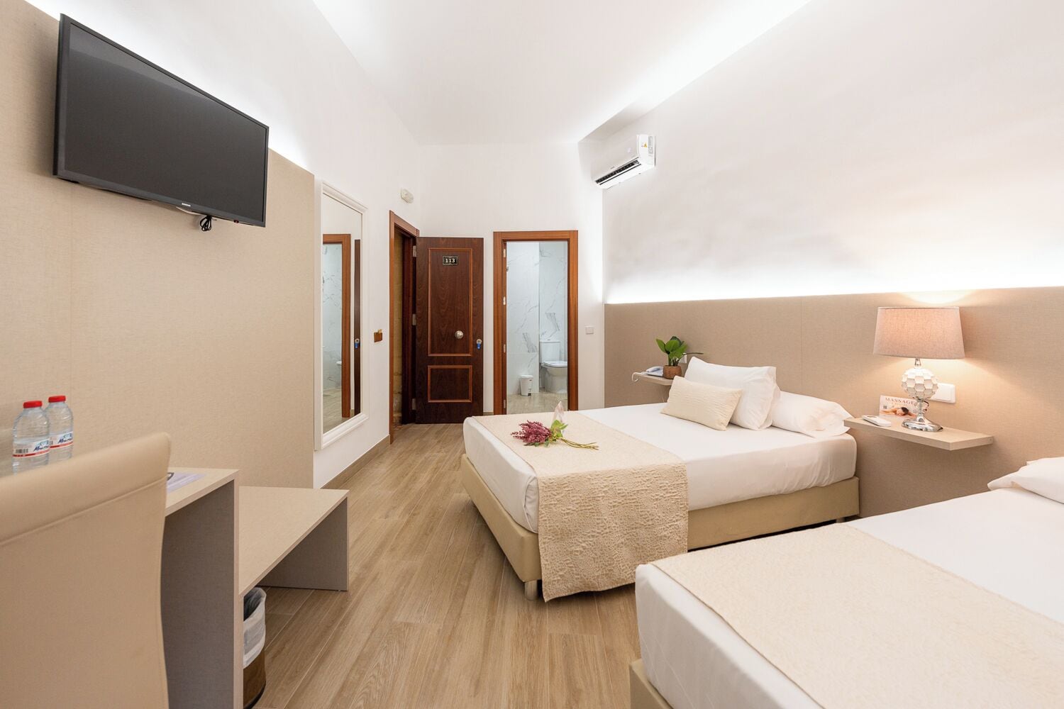 A hotel room with a bed, bedside table, lamp, and a TV mounted on the wall.