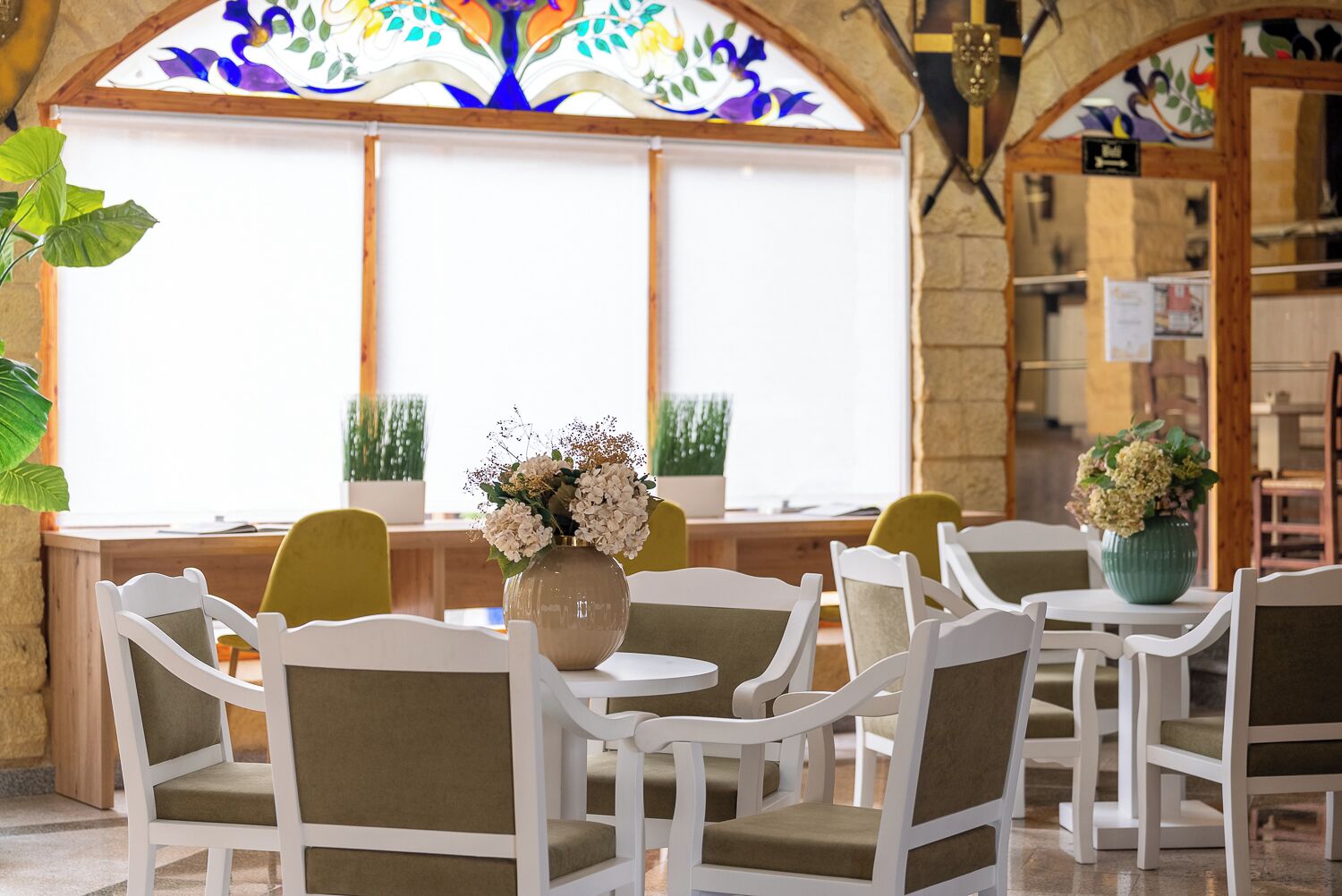 A dining area with white chairs and tables, a vase with flowers, and a stained glass window with a tree design.