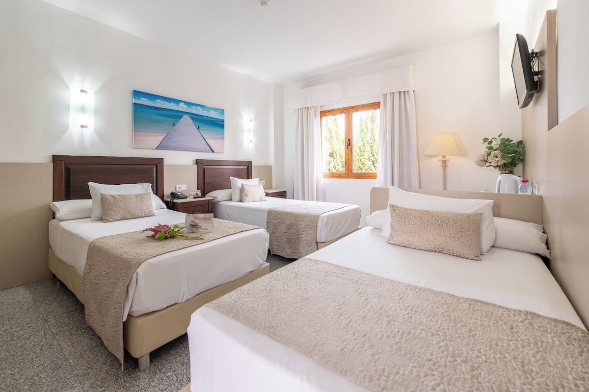 A hotel room with three beds, a painting of a beach, a window with curtains, and a wall-mounted TV.