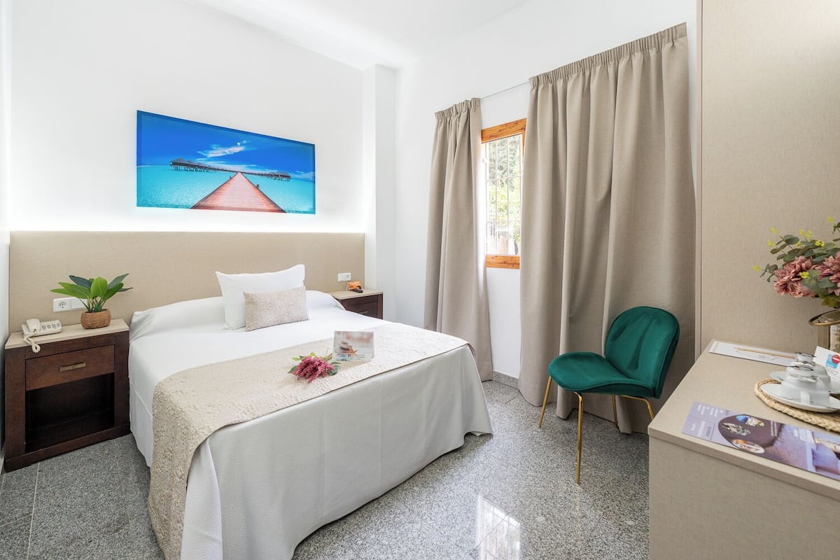 A hotel room with a bed, a desk, a chair, a nightstand, a phone, a plant, and a painting of a pier.