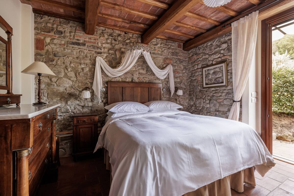 A bedroom with a stone wall, wooden ceiling beams, a bed with white linens, a nightstand, a dresser, and a window with curtains.