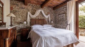 Quarto com parede de pedra, vigas de madeira no teto, cama com roupa de cama branca, mesa de cabeceira, cômoda e janela com cortinas.