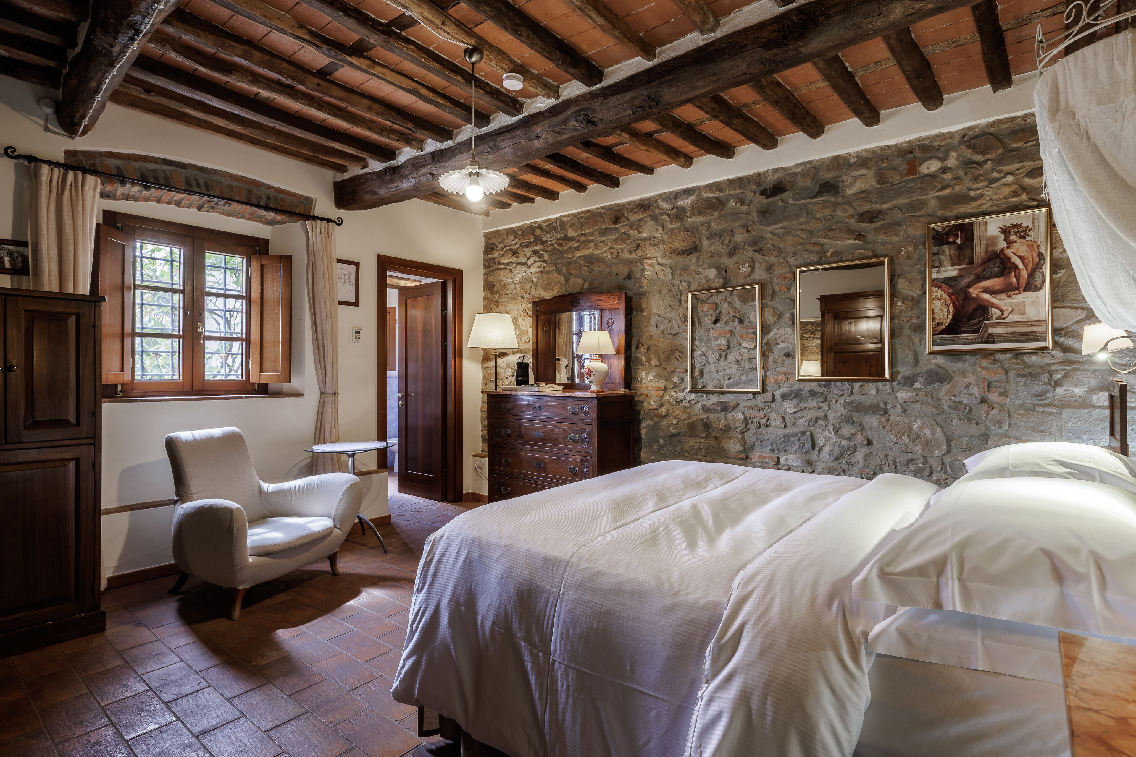 Een slaapkamer met een stenen muur, houten balken in het plafond, een bed, een stoel en een dressoir.