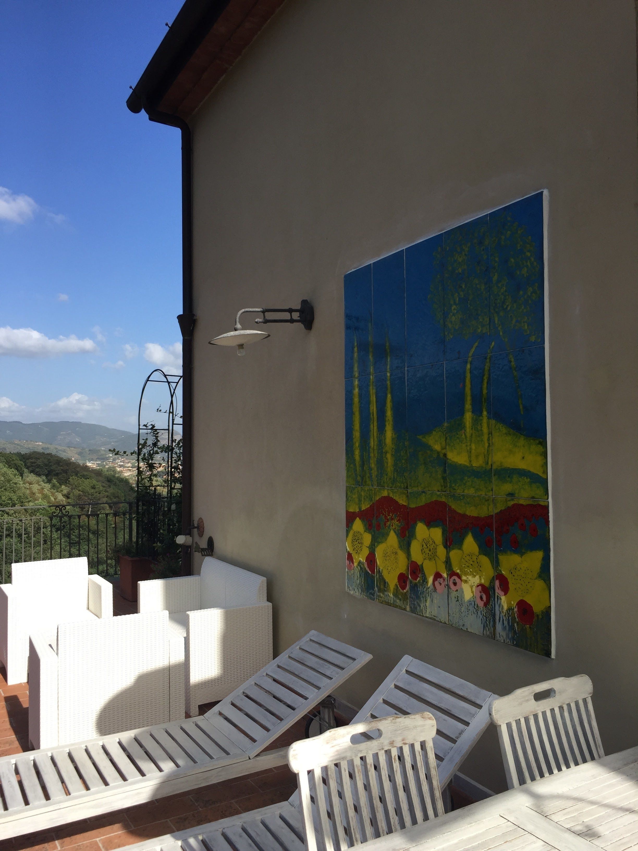 Eine Terrasse mit weißen Liegestühlen und einem Tisch, eine Wand mit einem farbenfrohen Wandgemälde und Blick auf die Landschaft.