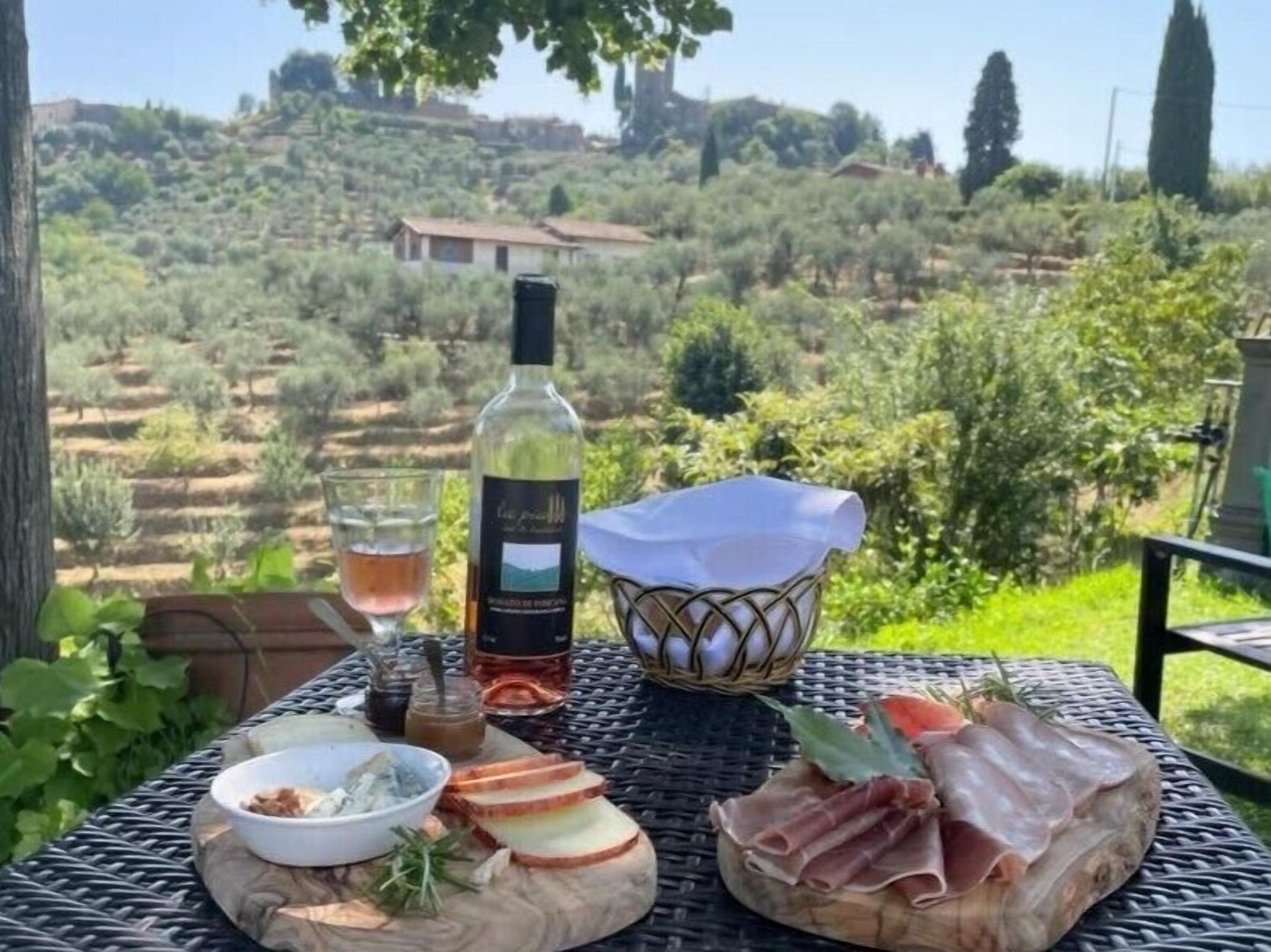 Uma mesa posta ao ar livre, com uma garrafa de vinho, um copo, uma cesta de ovos, uma tigela de azeitonas e fatias de carne curada.
