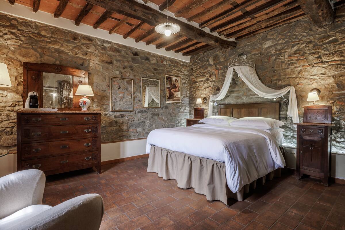 A bedroom with a stone wall, wooden ceiling beams, a four-poster bed, a dresser, and a chair.