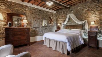 Une chambre avec un mur en pierre, des poutres apparentes en bois au plafond, un lit à baldaquin, une commode et un fauteuil.