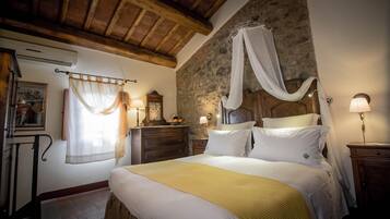 Une chambre avec un mur en pierre, un plafond en bois, un lit avec une literie blanche et jaune, une table de chevet avec une lampe et une commode en bois.