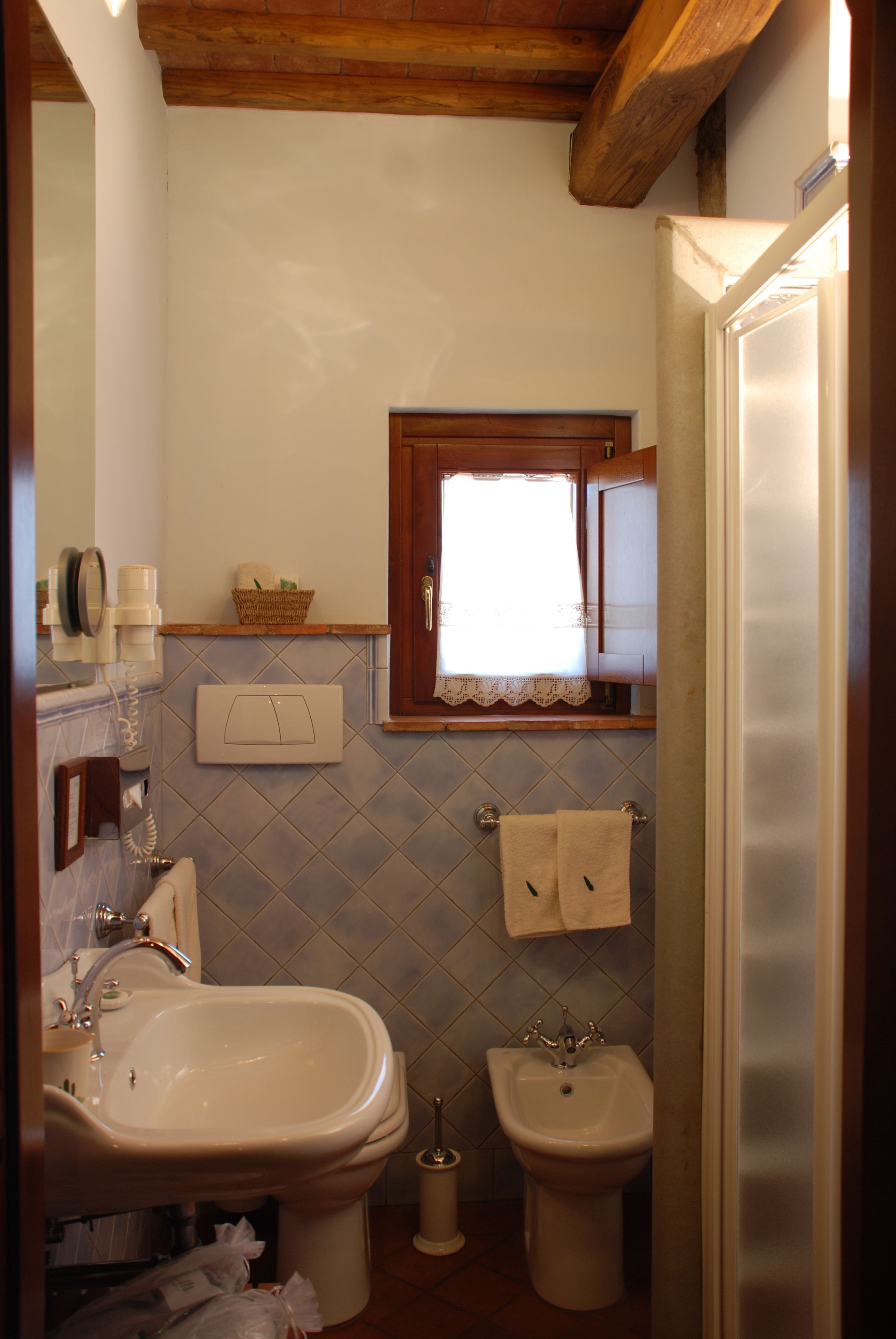 Ein Badezimmer mit Waschbecken, Bidet und Spiegel. Ein Fenster mit Spitze und ein Korb an der Wand sind ebenfalls vorhanden.