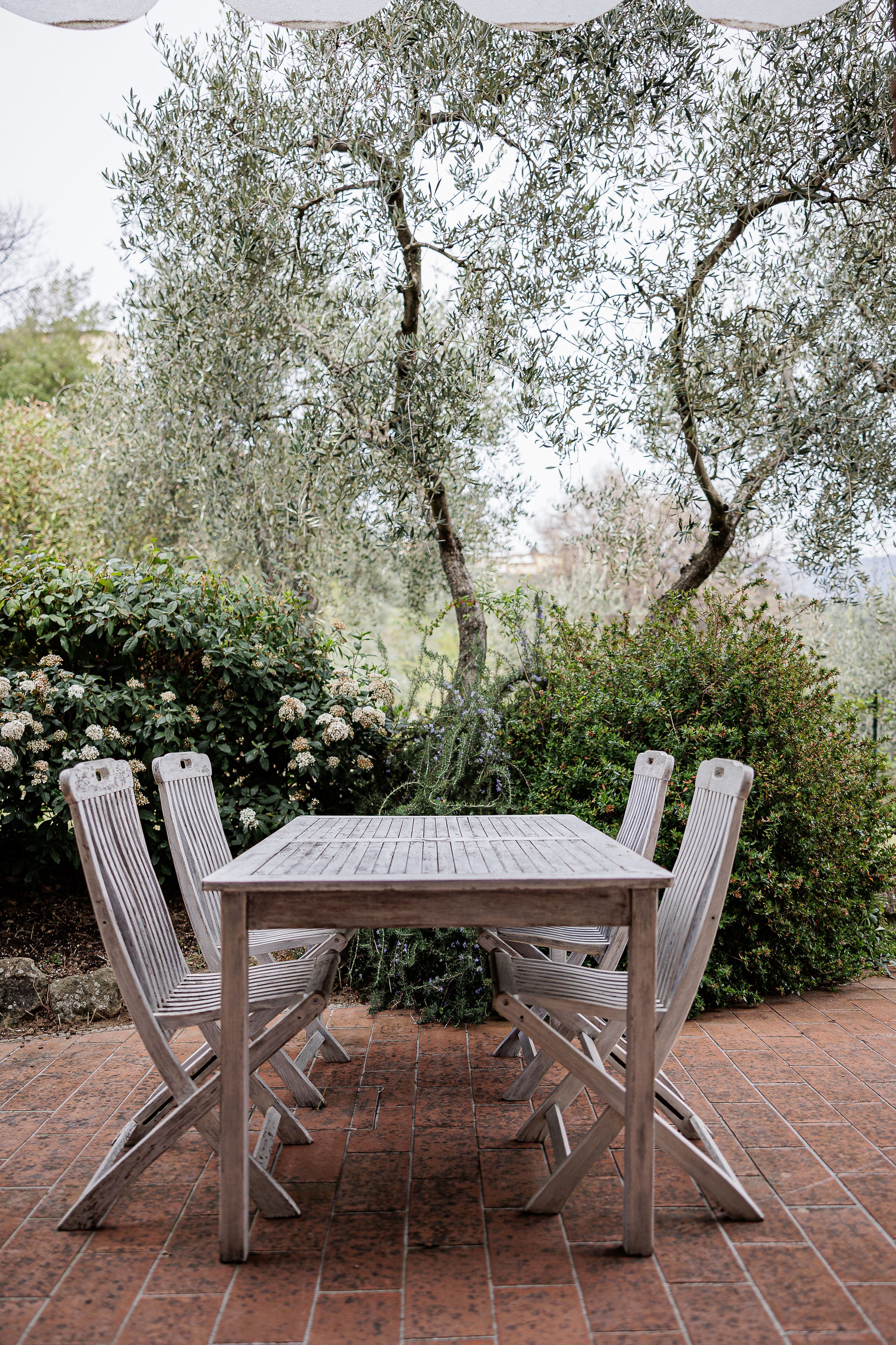 A wooden table and four chairs set outdoors on a tiled patio, surrounded by greenery and trees.