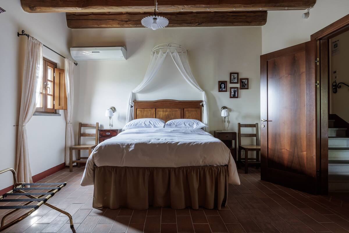 A bedroom with a wooden bed, a canopy, a wooden wardrobe, a chair, a nightstand, and a window with curtains.