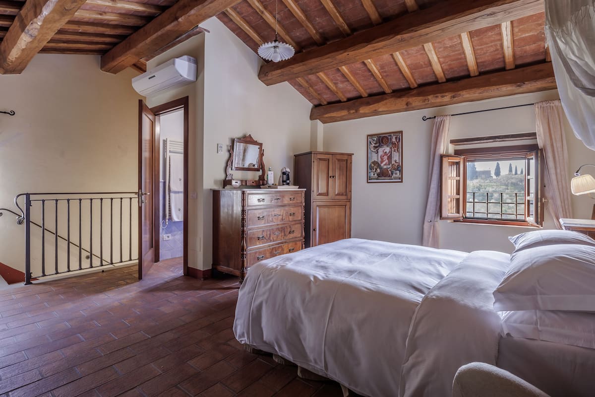 A bedroom with a wooden ceiling, a bed with white bedding, a dresser, a window with a view, and a door leading to another room.