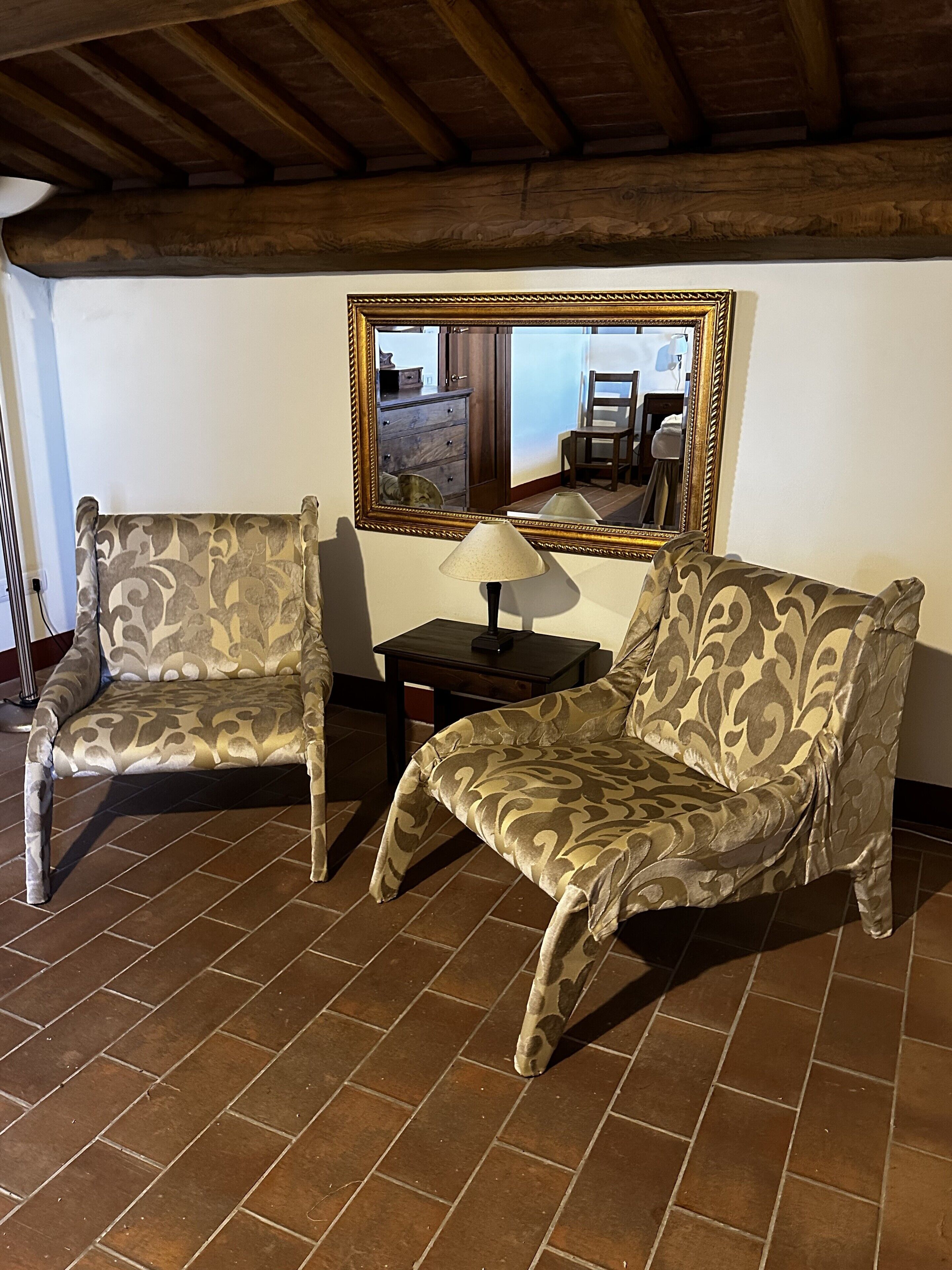 Una habitación con dos sillones estampados, una mesita con una lámpara y un espejo grande en la pared.