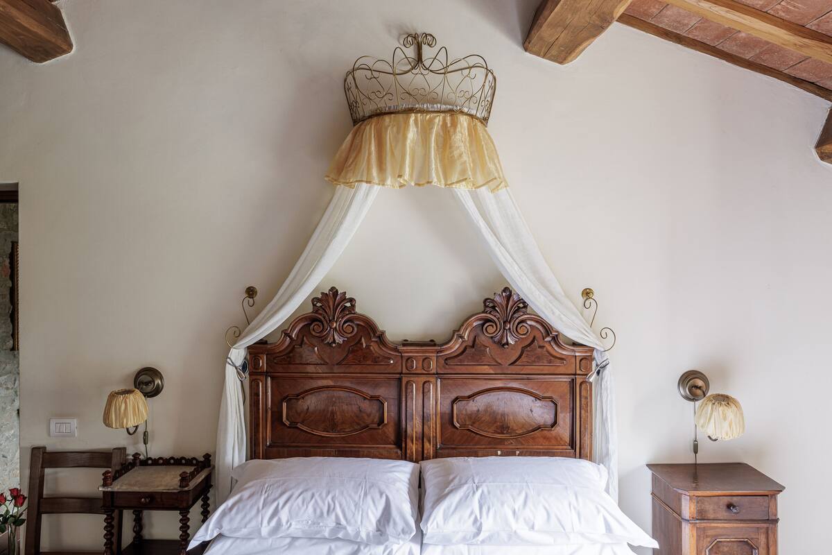 A four-poster bed with a canopy, a wooden nightstand with a lamp, and a chair with a vase of flowers.
