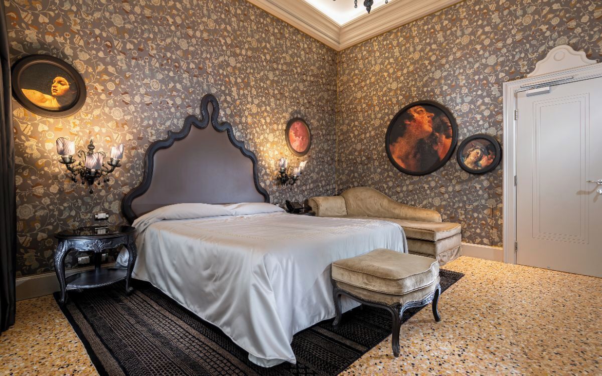 A bedroom with a large bed, a bedside table, a chair, and a wall with patterned wallpaper and framed pictures.
