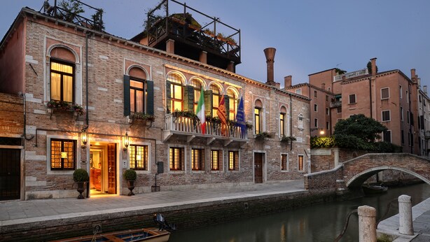 A historic building with a canal, a boat docked in front, and a bridge in the background.