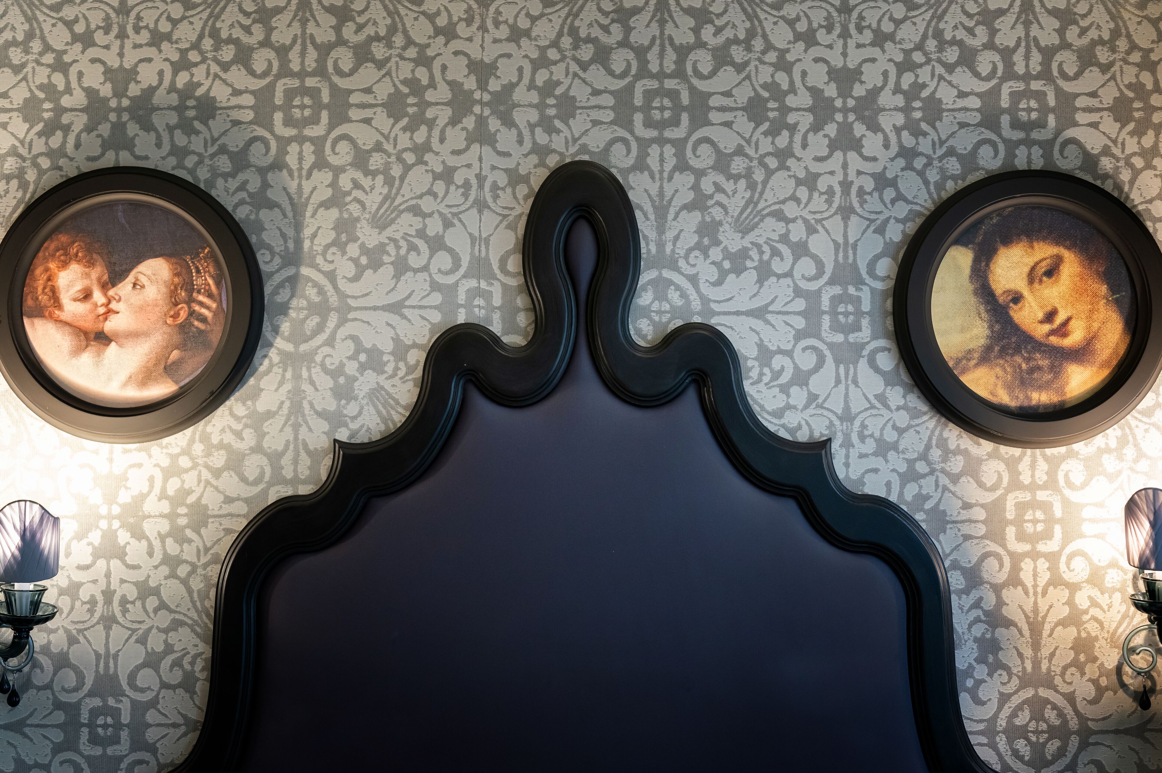 A room with a dark headboard, two wall-mounted picture frames, and a patterned wallpaper.