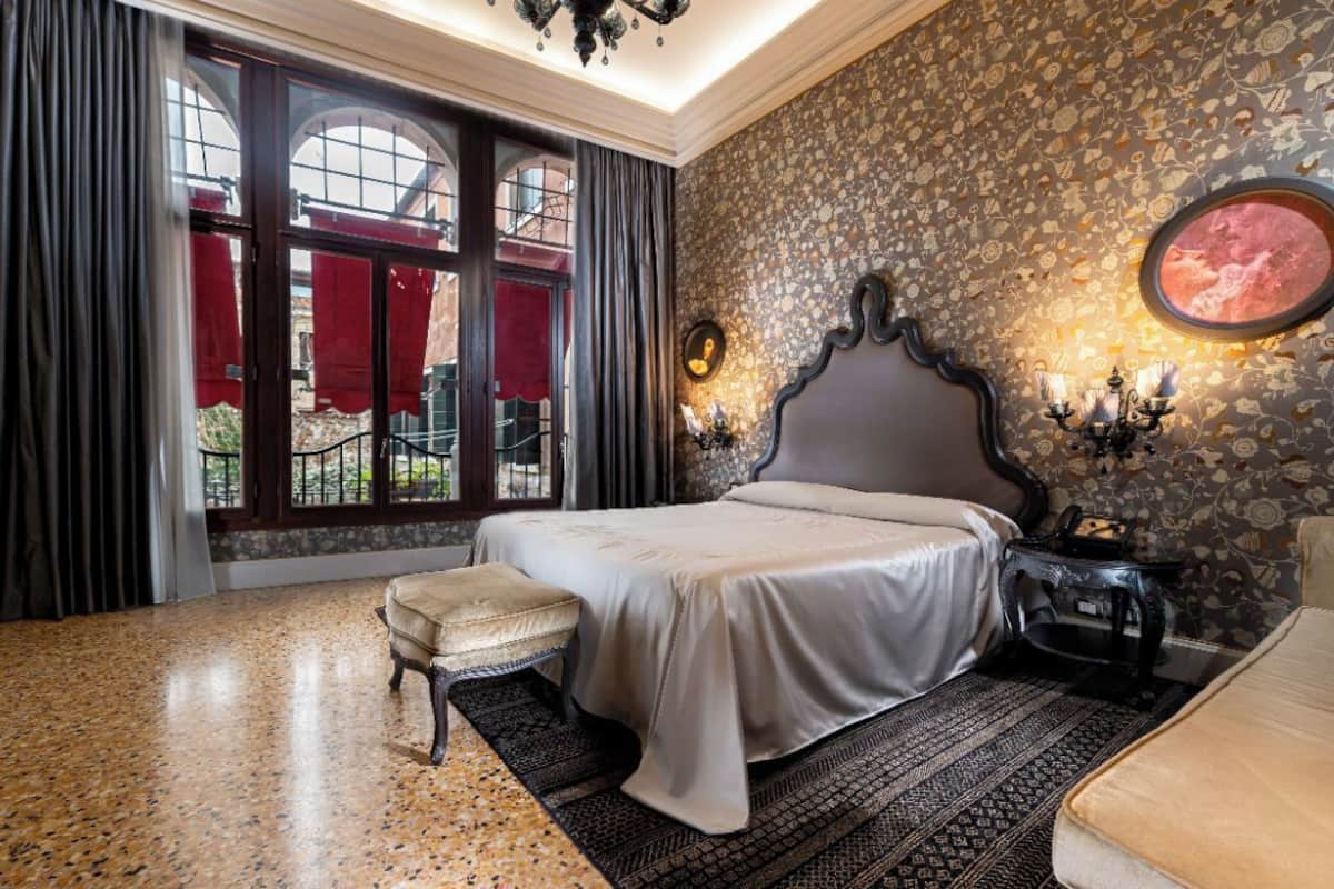A bedroom with a large bed, a bench, a nightstand with a lamp, and a patterned wall.