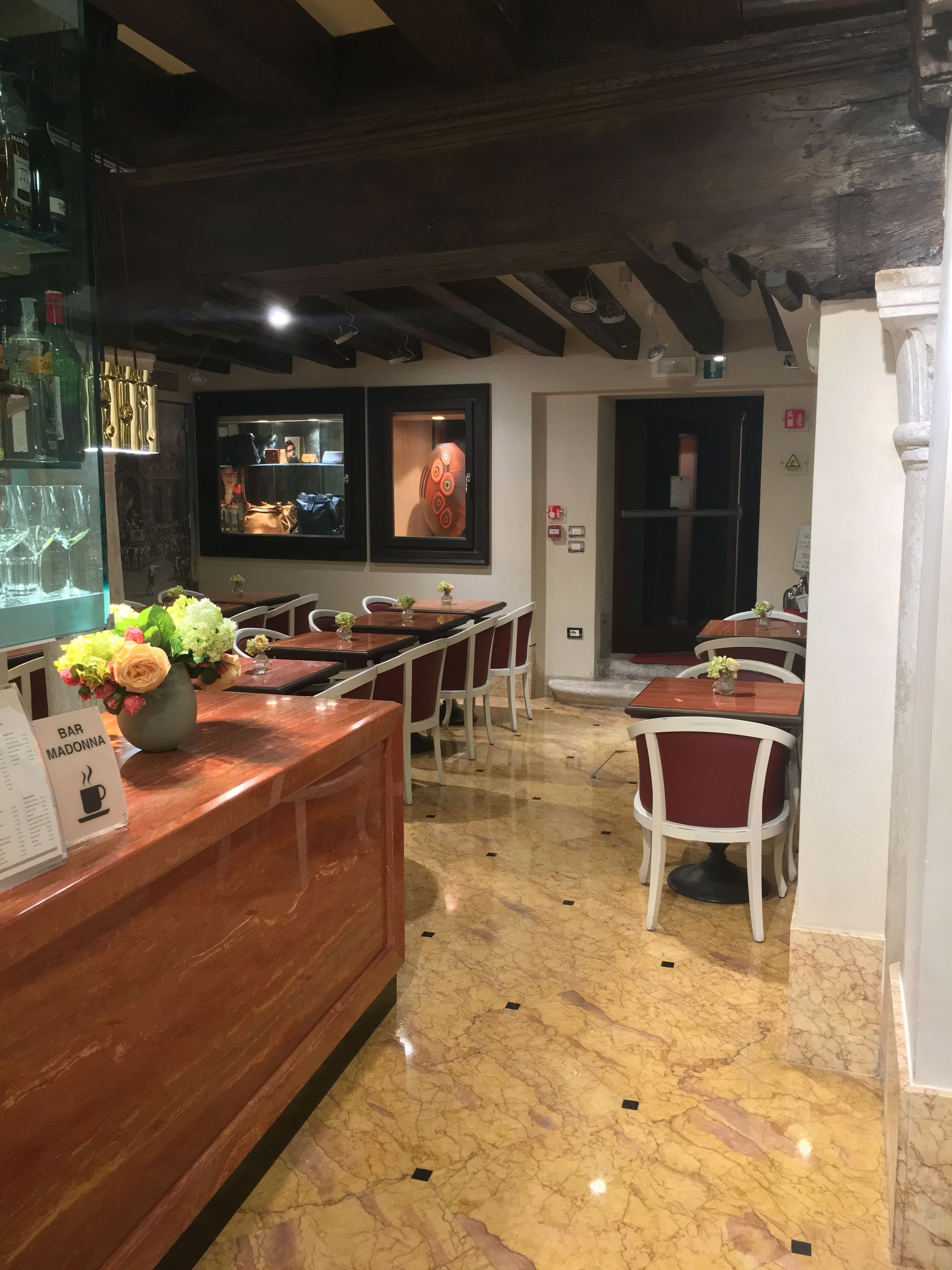 L’intérieur d’un restaurant présente un sol en marbre, des tables et des chaises en bois. On peut y admirer des compositions florales sur le comptoir et un téléviseur fixé au mur.