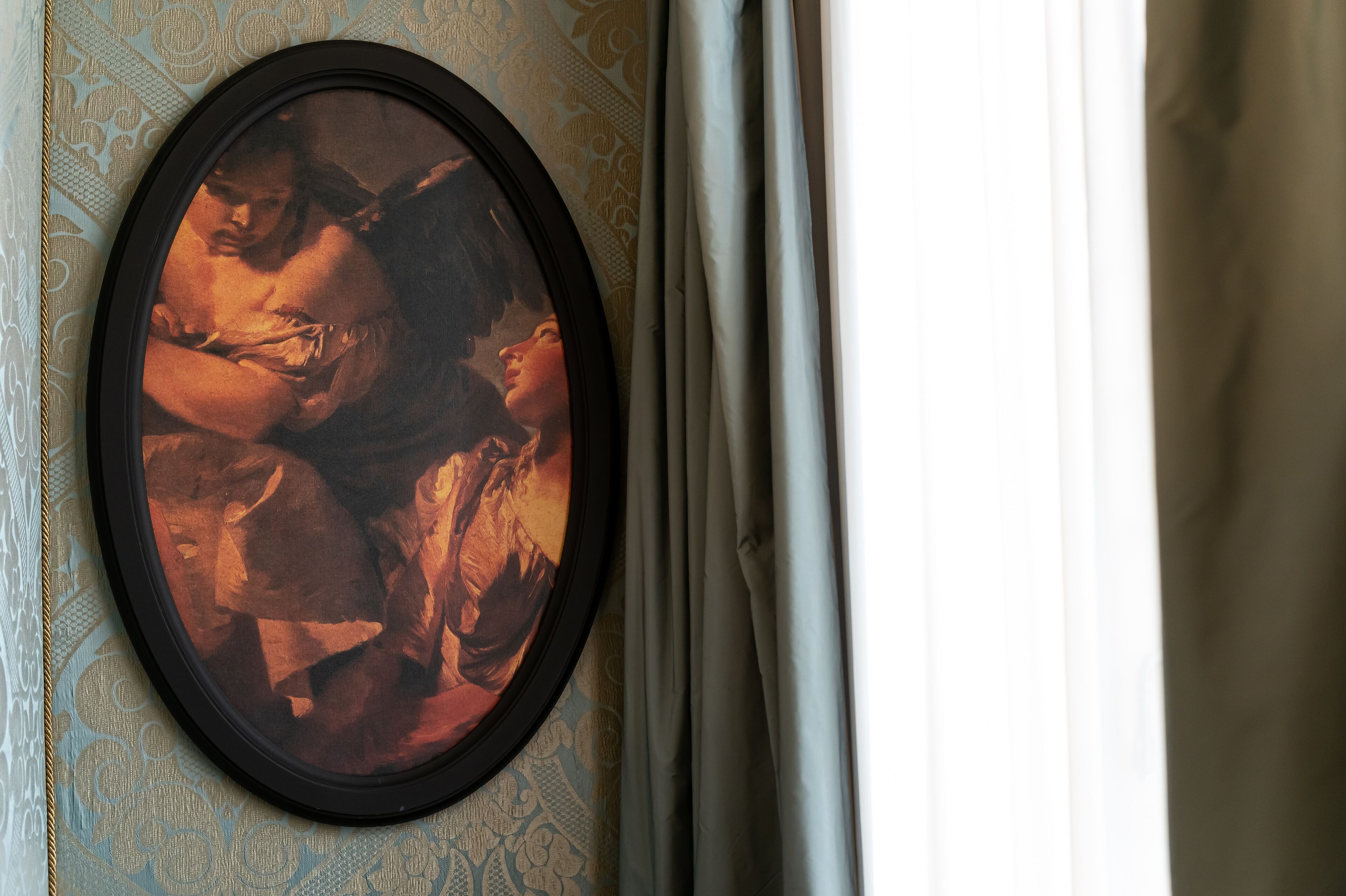 A framed artwork on a wall with a patterned wallpaper and a curtain partially drawn to the side.