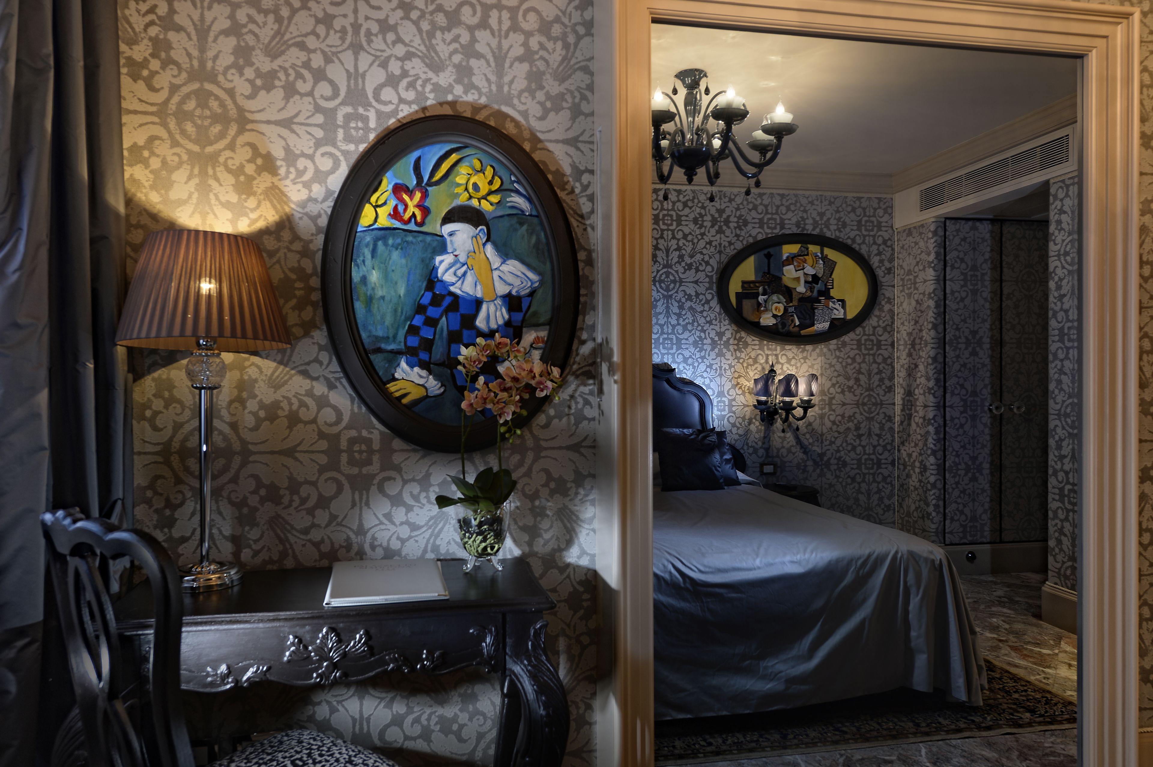A hotel room with a bed, a desk with a lamp, a mirror, and a wall with a decorative painting.