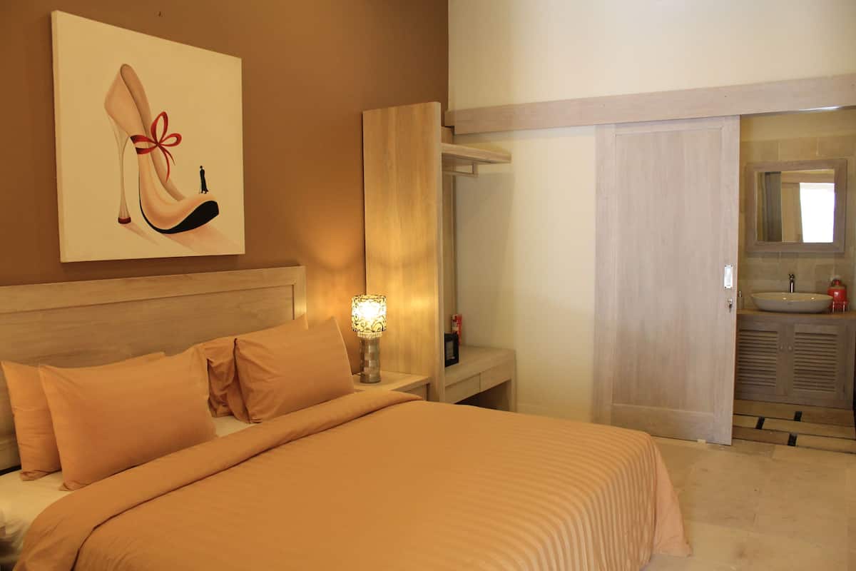 Habitación de hotel con cama, mesita de noche con lámpara, baño con lavamanos y espejo, y una puerta que da a otra habitación.
