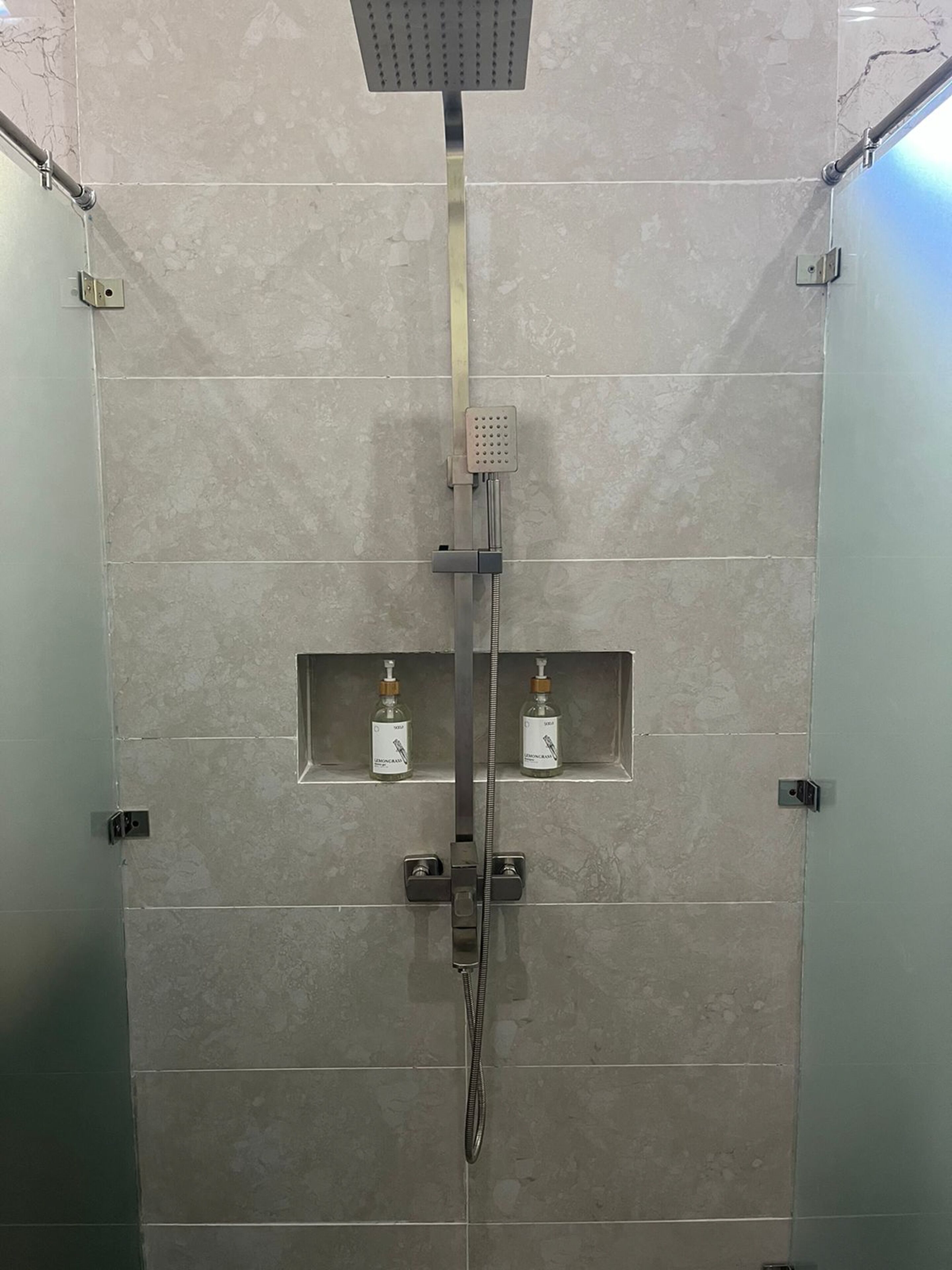 A modern shower with a handheld showerhead and a fixed rainhead, two small shelves with bottles, and a metal grab bar.
