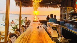Un bar offrant une vue sur le coucher de soleil sur l’eau, avec des meubles en bois et un toit de chaume.