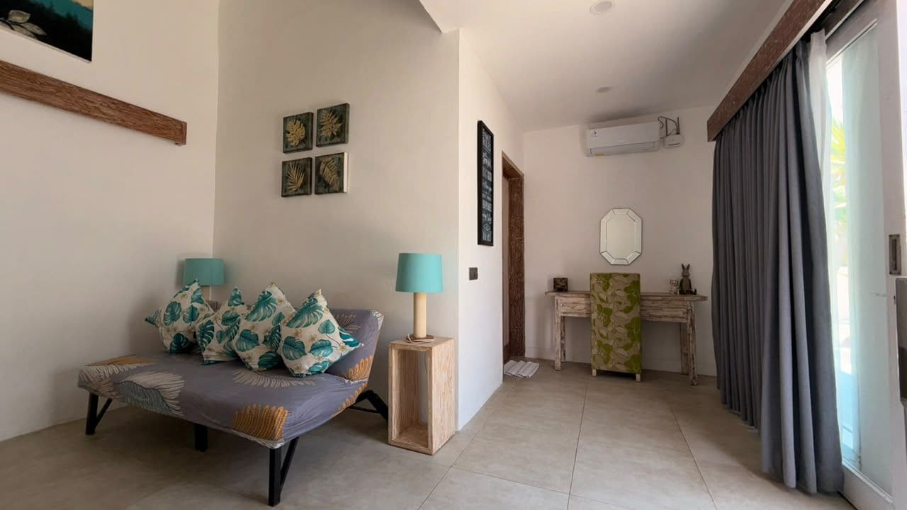 A modern living room with a sofa, a side table, and a lamp. There is a small table with a mirror and a decorative item. The room has a large window with curtains.